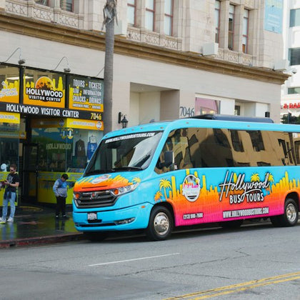 A bright blue Hollywood tour bus parked in front of a visitor center building with a sign reading 'Hollywood Visitor Center.'