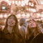 Two people look up and smile under warm, glowing lights in a festive indoor setting.