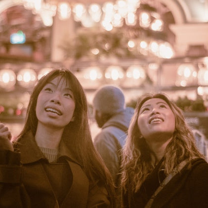 Two people look up and smile under warm, glowing lights in a festive indoor setting.