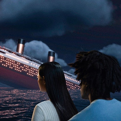 Two people watch a large, illuminated ship sinking in the night ocean under cloudy skies.