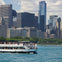 A boat named 'Lila' with passengers on a river, with a city skyline featuring tall buildings and greenery in the background.