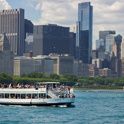 A boat named 'Lila' with passengers on a river, with a city skyline featuring tall buildings and greenery in the background.