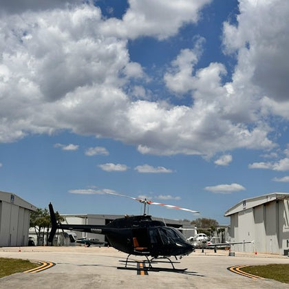 A black helicopter sits on a runway between two hangars under a cloudy blue sky.