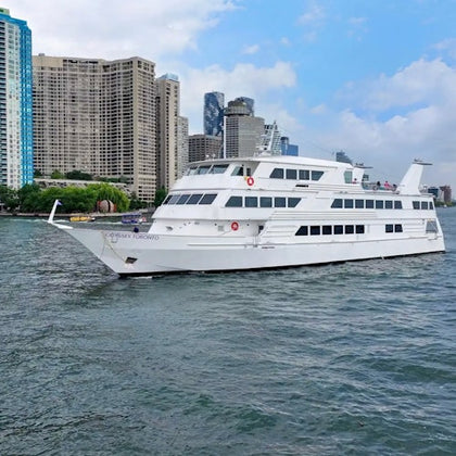 A large white passenger boat on a city river, with tall buildings and a blue sky in the background.