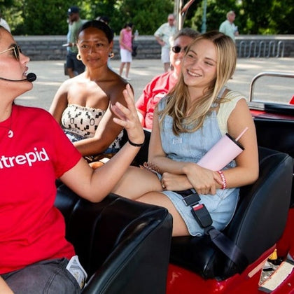 A group of people seated on a tour vehicle, with one wearing a microphone and speaking, and others smiling and listening.