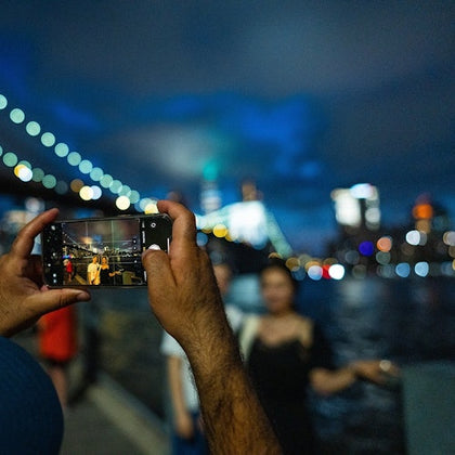 A person is taking a photo with a smartphone of two other people posing near a body of water at night, with city lights in the background.