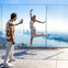 A man takes a photo of a woman posing playfully on a glass observation deck overlooking a city and waterfront.