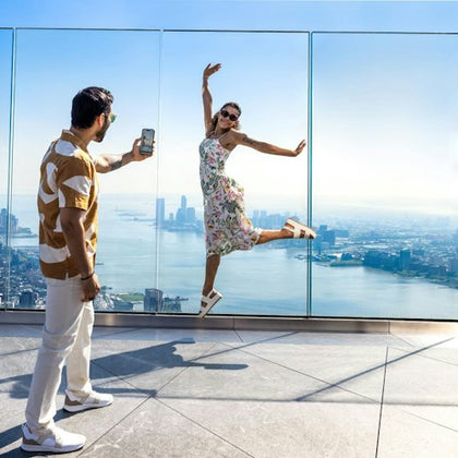 A man photographs a woman posing with a jump in front of a glass barrier overlooking a cityscape and body of water.