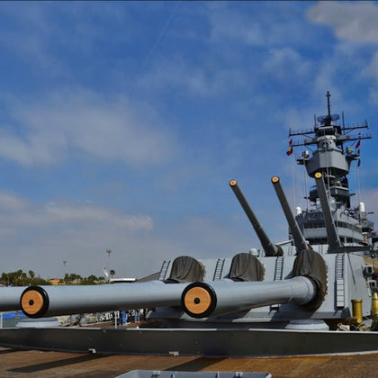 Large naval battleship with three gun turrets in the foreground, docked under a blue sky with light clouds.
