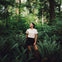 A person stands in a lush forest surrounded by ferns, wearing a white shirt and black shorts, looking upward.