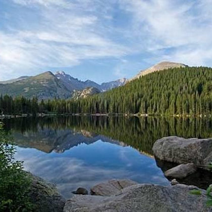 A serene mountain lake with clear reflections of the tree-lined shore and rugged peaks under a partly cloudy sky.