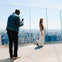 A man photographs a smiling woman standing on a glass-fenced observation deck overlooking a city skyline.