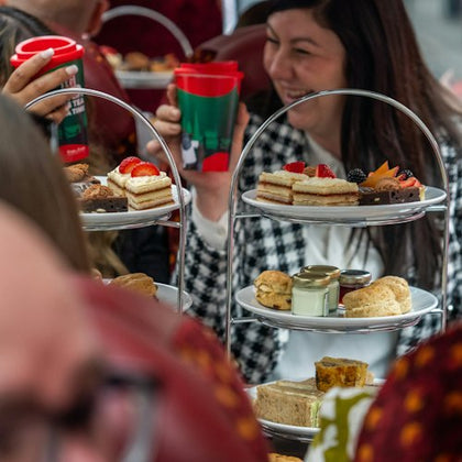 People enjoying an afternoon tea with tiered trays of cakes and sandwiches, holding festive red cups, smiling.