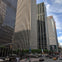 City street with tall office buildings, pedestrians, cars, and taxis under a partly cloudy sky.