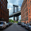 A street view framed by tall red brick buildings with the Manhattan Bridge in the background under a partly cloudy sky.