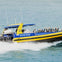 A bright yellow speedboat passes by a waterfront building with flags waving, under a clear blue sky.