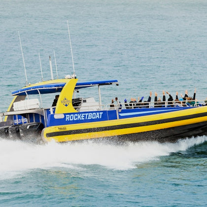 A bright yellow speedboat passes by a waterfront building with flags waving, under a clear blue sky.