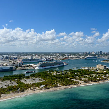 Aerial view of a coastal city with docks, multiple cruise ships, and urban buildings under a partly cloudy sky.