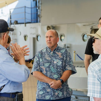 A man in a patterned shirt stands on a ship's deck, surrounded by others listening to a discussion.