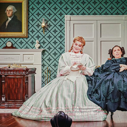 Two people in period attire sit in a historical room with patterned walls, a fireplace, and a portrait of George Washington.
