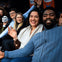 A group of smiling people sitting together, waving at the camera, with a person holding a skull prop on the left.