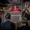 A man in red robes and cap stands in a wooden pulpit, addressing three onlookers. Shelves with books are visible in the background.
