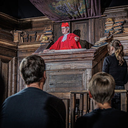 A man in red robes and cap stands in a wooden pulpit, addressing three onlookers. Shelves with books are visible in the background.