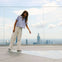 A woman on a rooftop observation deck pointing at the ground, with a city skyline and glass barriers in the background.