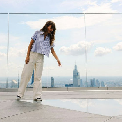A woman on a rooftop observation deck pointing at the ground, with a city skyline and glass barriers in the background.