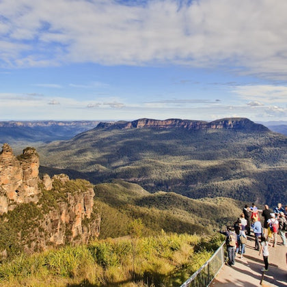 A scenic view of a forested valley with cliffs, under a partly cloudy sky, and a group of tourists on a viewing platform.