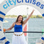 A woman in a blue top and white skirt stands on a boat, smiling and raising her arm, with water and trees in the background.