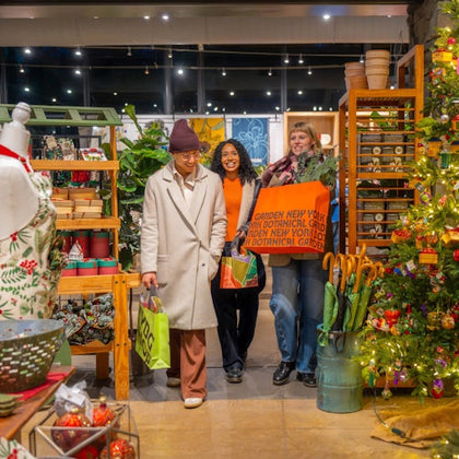 Three people holding shopping bags in a colorful store decorated with plants, shelves, and a lit Christmas tree.