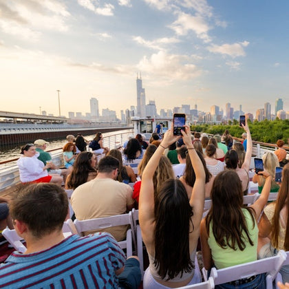 A group of people seated outdoors on a boat, facing a city skyline, with some holding up phones to take photos.