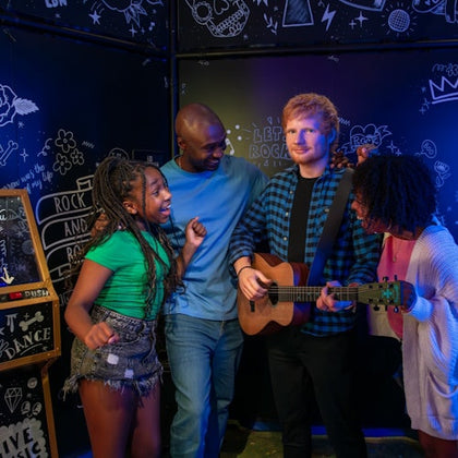 Four people stand together in a music-themed room; one holds a guitar while the others smile and gesture excitedly.