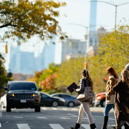 People crossing a city street at a crosswalk, with trees and parked cars in the background.