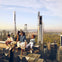 Four people sitting on a steel beam high above a cityscape with skyscrapers and a park visible in the background.