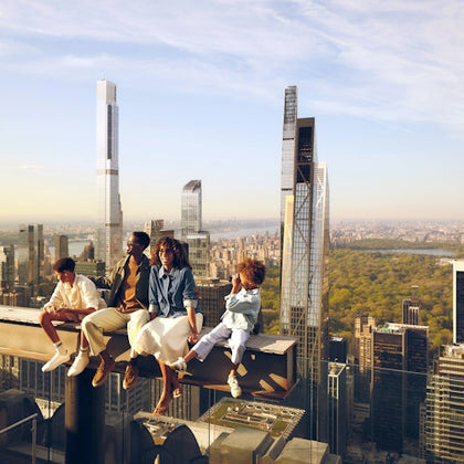 Four people sitting on a steel beam high above a cityscape with skyscrapers and a park visible in the background.
