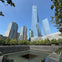 A memorial pool surrounded by trees with modern skyscrapers, including One World Trade Center, under a clear blue sky.