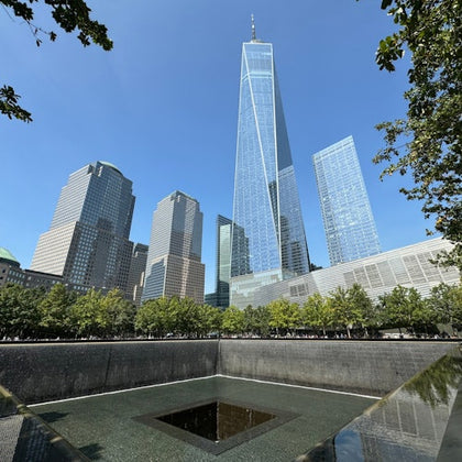 A memorial pool surrounded by trees with modern skyscrapers, including One World Trade Center, under a clear blue sky.