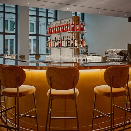 A modern bar with a curved counter, upholstered stools, and shelves displaying various liquor bottles under soft lighting.