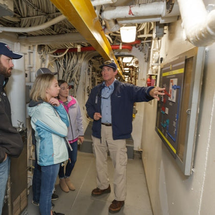 A tour guide explains a diagram to a small group of people in an industrial setting with pipes and equipment visible.