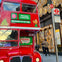 A red double-decker bus promoting an afternoon tea tour is parked near a decorated building and a bus stop in an urban area.