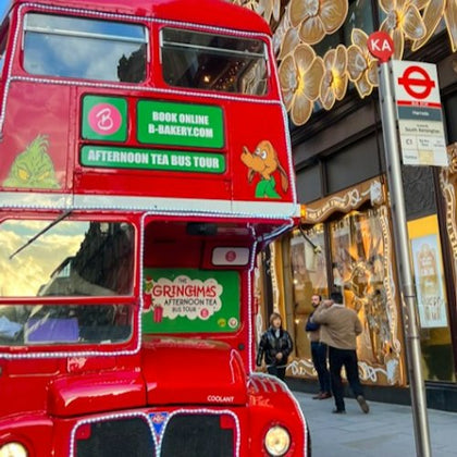 A red double-decker bus promoting an afternoon tea tour is parked near a decorated building and a bus stop in an urban area.