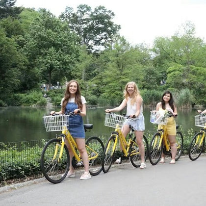 Three women standing next to yellow bicycles beside a pond in a park with lush greenery.