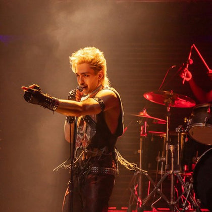 A performer with blonde hair sings into a microphone on stage with a drum set in the background under dramatic lighting.