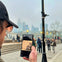 A person holding a phone displaying a digital artwork, with a cityscape and people walking in a foggy urban setting.