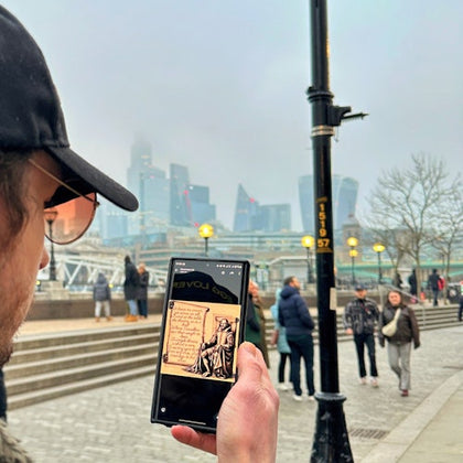 A person holding a phone displaying a digital artwork, with a cityscape and people walking in a foggy urban setting.