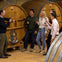Five people standing and holding wine glasses, conversing in a winery with large wooden wine barrels in the background.