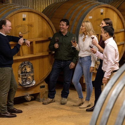 Five people standing and holding wine glasses, conversing in a winery with large wooden wine barrels in the background.