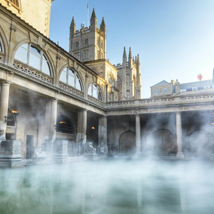 A historic bathhouse with steamy water, surrounded by columns and arches, with a cathedral visible in the background.